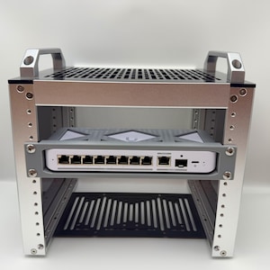 以下が含まれることがあります： 黒い上部と下部の棚が付いたシルバーの金属製ラック。中央の灰色の棚に灰色のデバイスが取り付けられています。デバイスには複数のポートと白いフロントパネルがあります。ラックには2つのシルバーのハンドルが付いています。