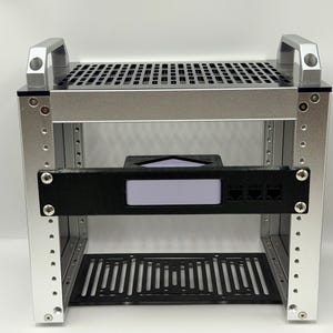Puede incluir: Un bastidor de servidor de marco abierto de metal plateado con una rejilla de ventilación superior y dos asas. Un soporte de montaje horizontal negro está fijado en la parte frontal, con una abertura rectangular central y tres puertos circulares.