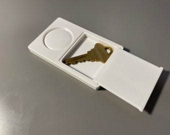 May include: A white plastic key holder with a hinged lid. The lid is open and a gold key is inside.
