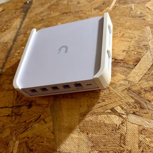 May include: A white electronic device with a curved top and a small, grey 'U' logo. The device has several ports on the front and is mounted in a white frame. The background is a textured, brown surface.