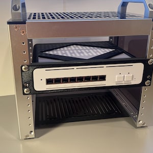Op de afbeelding: Een zilveren metalen serverrack met een zwart-wit netwerkapparaat erin gemonteerd. Het rack heeft een geperforeerde boven- en onderplank, met een handvat aan de bovenkant. Het netwerkapparaat heeft meerdere poorten en knoppen.
