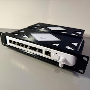 Op de afbeelding: Zwart-wit netwerkapparaat met meerdere poorten, gemonteerd in een rek. Het apparaat heeft een witte behuizing met een zwarte, geometrisch gevormde afdekking. Het beschikt over een kleine antenne en diverse aansluitpoorten.