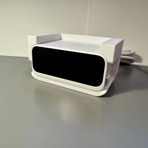 May include: A white electronic device with a black screen, rectangular in shape, and a rounded front. It has a white power cord and plug. The device is on a gray surface.