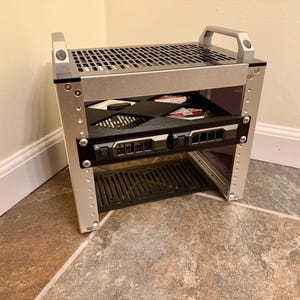 May include: A silver metal rack with a handle on top and multiple shelves. The rack has a perforated top and a black shelf with various electronic components. The rack is designed to hold electronic devices and has a sturdy, industrial appearance.