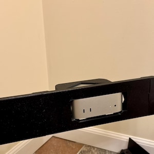 May include: Black metal rack mount with a silver computer component installed. The rack has multiple screw holes and is mounted against a cream wall. The computer component has two vertical slots.