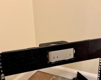 May include: Black metal rack mount with a silver computer component installed. The rack has multiple screw holes and is mounted against a beige wall. The computer component has two vertical slots.