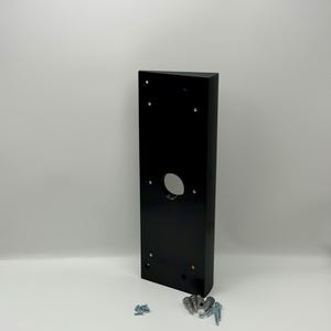May include: A rectangular black doorbell mount with a circular opening and several screw holes. A set of screws and anchors are placed below the mount. The mount is made of a glossy material.