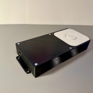 May include: A black rectangular electronic device with a white top section. The device has a port and is secured with small silver screws. The top section has a circular button. The device is on a gray surface.