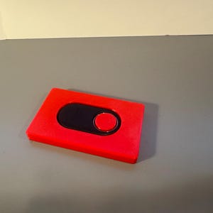 May include: A red rectangular object with a black oval and a red circle. The object is on a gray surface. The red color is bright and the object appears to be made of plastic.