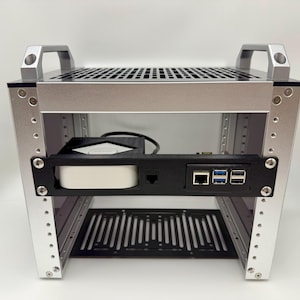 May include: Silver and black computer case with a perforated top and bottom. The case has a black panel with ports and a white device. Two silver handles are on top. The case is designed to hold computer components.