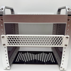 Pode incluir: Um rack de metal prateado com um painel de malha de favo de mel branco e um painel ranhurado preto. O rack tem duas alças na parte superior e foi projetado para conter equipamentos. O rack tem aproximadamente 48 cm de largura.
