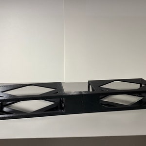 Könnte beinhalten: Schwarzer Metallständer mit zwei offenen Fächern, entworfen, um elektronische Geräte zu halten. Der Ständer hat ein geometrisches Muster und ist aus robustem Metall gefertigt.