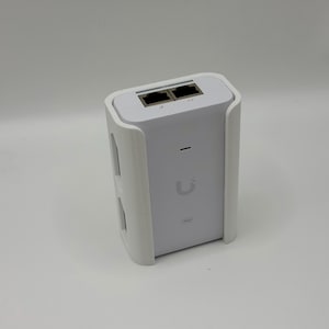 May include: A white network device featuring a rectangular body with two Ethernet ports on the top surface. The front panel displays a small horizontal status light, a central U-shaped logo, and a small rectangular indicator near the bottom edge.