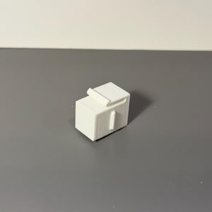 Peut inclure: Un petit objet blanc imprimé en 3D avec une base rectangulaire et un dessus incurvé en relief. Une barre verticale s'étend de la base. L'objet est sur une surface grise.