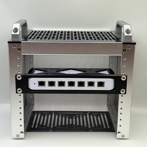 May include: A silver metal rack with a black base and top, designed to hold electronic equipment. The rack has a handle on top and a black panel with multiple ports. The rack is designed to hold electronic equipment.