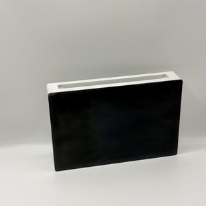 May include: A rectangular, flat-sided container featuring a solid black front panel and a contrasting white frame. The top edge of the object has a long, narrow opening that runs across the entire length of the structure, creating a sleek, open-top design.