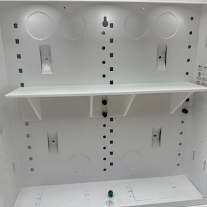 May include: A white, rectangular enclosure with a shelf and various circular and square openings. The interior features mounting points and a green button. A label with text and a QR code is visible on the right side. The enclosure is designed for electrical or network equipment.
