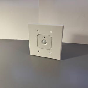 Puede incluir: Un sensor de movimiento blanco, de forma cuadrada, con un cuadrado más pequeño y empotrado en el centro. Cuenta con una lente redondeada y está montado en una placa de pared blanca con cuatro orificios para tornillos. El fondo es una superficie gris y una pared blanca.