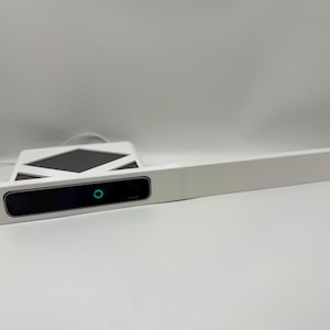 May include: A white rack-mount panel featuring a central black rectangular display module with a small, glowing teal circular indicator light. The panel has mounting holes on the left side and an extended flat white horizontal surface section.