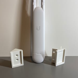 Könnte beinhalten: Ein weißer Ubiquiti-Zugangspunkt mit zwei Antennen, flankiert von zwei weißen 3D-gedruckten Montagehalterungen. Auf den Halterungen steht der Text "3DEDSTUFF". Der Zugangspunkt hat das Ubiquiti-Logo.