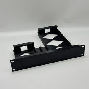 May include: A black 3D-printed rack mount shelf featuring a solid front panel with mounting holes on each side. The structure includes two distinct recessed compartments with open diamond-shaped cutouts for ventilation and equipment support.