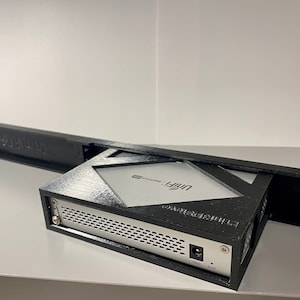 May include: A black 3D printed rack mount enclosure for a UniFi Dream Machine Pro network device. The enclosure has a black plastic front panel with a cut-out for the device's front panel and a black plastic rear panel with a cut-out for the device's rear panel. The enclosure is designed to be mounted in a standard 19-inch rack.