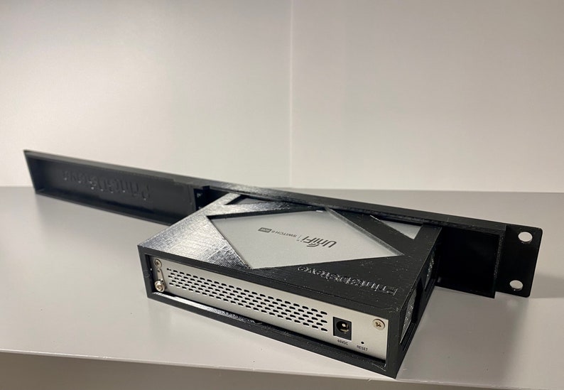 May include: A black 3D printed rack mount enclosure for a UniFi Dream Machine Pro network device. The enclosure has a black plastic front panel with a cut-out for the device's front panel and a black plastic rear panel with a cut-out for the device's rear panel. The enclosure is designed to be mounted in a standard 19-inch rack.