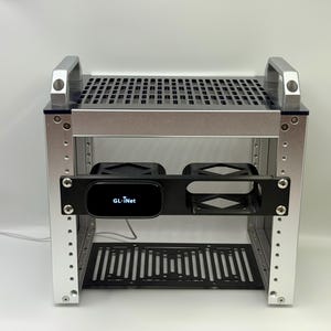 Op de afbeelding: Een zilveren metalen rek met een zwart geperforeerde boven- en onderplank. Een zwart paneel in het midden bevat twee compartimenten en een apparaat met de tekst "GL-Inet". Het rek heeft zilveren handgrepen aan de bovenkant.