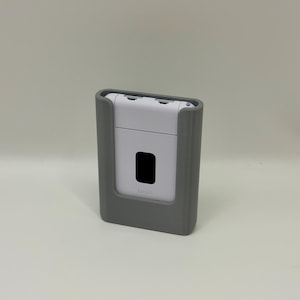 Może przedstawiać: Szare i białe urządzenie elektroniczne o prostokątnym kształcie. Urządzenie posiada czarny prostokątny ekran i jest zamknięte w szarej obudowie. Urządzenie znajduje się na białej powierzchni.
