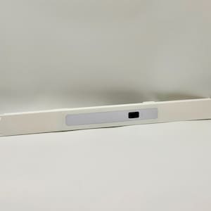 Puede incluir: Un panel de montaje en rack blanco, largo y estrecho, que presenta un recorte rectangular centrado con una inserción de color claro y una pequeña ventana rectangular oscura para sensor o indicador situada en el lado derecho de la inserción.