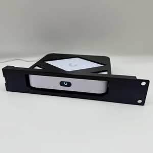 Può includere: Un supporto per rack nero con un dispositivo bianco e grigio. Il dispositivo ha un piccolo logo blu a forma di U illuminato. Il supporto per rack ha due fori di montaggio e un design geometrico.