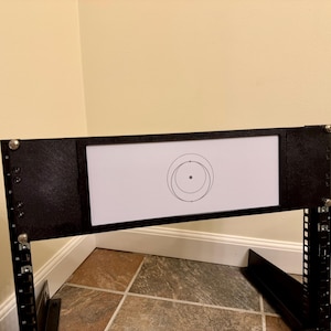 May include: Black metal frame with a white rectangular center. The white center features a black concentric circle design. The frame is supported by black metal legs and is mounted on a tiled floor.