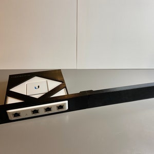 May include: A black plastic wall mount for a Ubiquiti UniFi network device. The mount has a rectangular base with four Ethernet ports and a slot for the device. The mount is attached to a long black plastic rail.