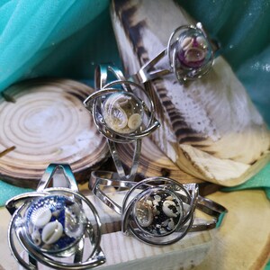 Peut inclure: Bracelets-manchettes en métal argenté avec un centre sphérique transparent rempli de coquillages et de sable. Les bracelets ont un design tourbillonnant unique autour de la sphère. Les bracelets sont exposés sur une surface en bois.