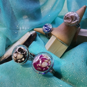 Peut inclure: Quatre bagues artisanales avec des globes en verre transparent, chacune contenant des éléments uniques. L'une présente du sable noir et de petites pierres, une autre des paillettes roses et des pierres, et deux autres du sable et des coquillages. Les bagues sont exposées sur un présentoir en bois.