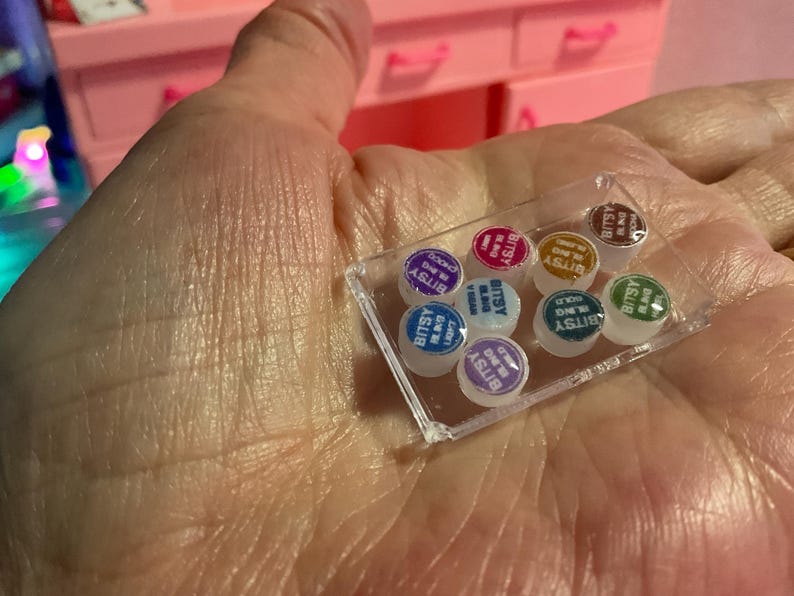 May include: A clear rectangular plastic tray containing eight miniature round containers. Each container features a colored lid with the text Bitsy Bling printed on it. The colors include purple, pink, gold, brown, blue, white, teal, and green.