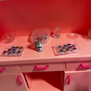 May include: A bright pink toy kitchen unit featuring a central miniature coffee cup labeled Bling Cafe, flanked by two clear plastic trays containing small colorful circular containers, a clear bowl, a dome-shaped item, and a small plate.