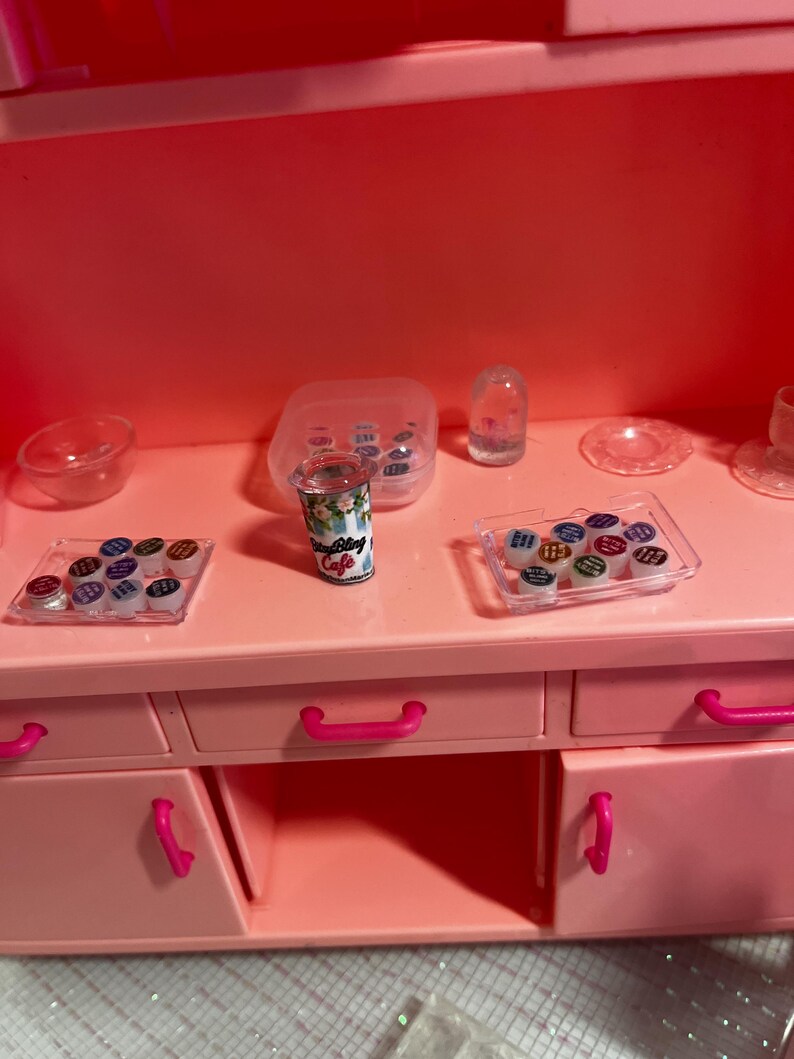 May include: A bright pink toy kitchen unit featuring a central miniature coffee cup labeled Bling Cafe, flanked by two clear plastic trays containing small colorful circular containers, a clear bowl, a dome-shaped item, and a small plate.