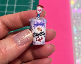 May include: A miniature plastic cup with a lid and straw, featuring a purple bow, a gold crown, and the text Bitsy Bling Café. A small topper with a pink heart sits on the blue straw. The cup has a white and purple watercolor-style background.
