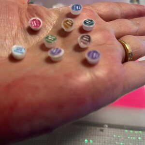 May include: A collection of nine small, circular, white-based containers with colored lids in shades of blue, pink, gold, teal, green, purple, and brown. Each lid features printed text, and the containers are arranged in a cluster on a surface.