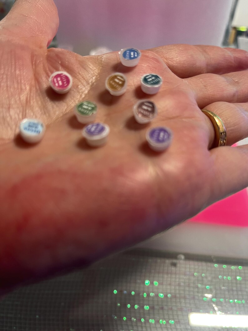 May include: A collection of nine small, circular, white-based containers with colored lids in shades of blue, pink, gold, teal, green, purple, and brown. Each lid features printed text, and the containers are arranged in a cluster on a surface.
