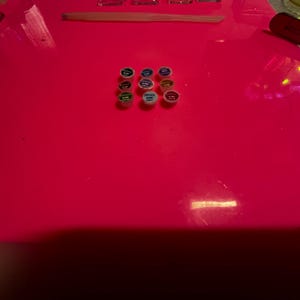 May include: A collection of nine small, circular containers arranged in a grid on a bright neon pink surface. Each container features a colored lid with text, including shades of green, blue, yellow, and red, alongside a tool and tray.