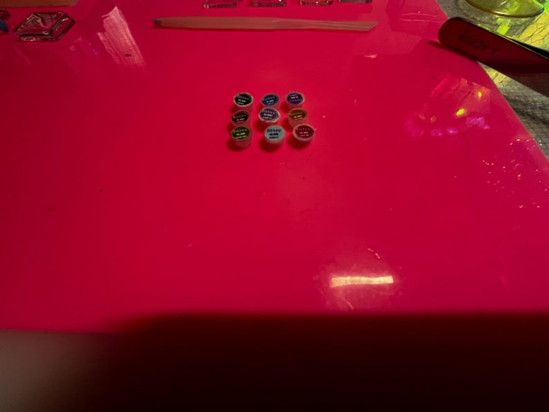 May include: A collection of nine small, circular containers arranged in a grid on a bright neon pink surface. Each container features a colored lid with text, including shades of green, blue, yellow, and red, alongside a tool and tray.