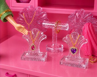 May include: Two clear acrylic shell-shaped jewelry stands displaying gold-tone necklaces with pink teardrop and purple heart pendants. A clear T-bar stand holds a pink stone ring. The items are set against a bright pink background with a doll arm.