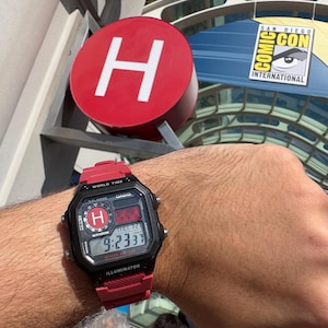 Könnte beinhalten: Eine rot-schwarze Digitalarmbanduhr mit quadratischem Zifferblatt, Weltzeitanzeige, Alarm und Illuminator-Funktion. Die Uhr hat ein rotes Kunstharzarmband und eine schwarze Lünette mit weißem Text, die die Uhrzeit 9:23 und den Buchstaben H anzeigt.