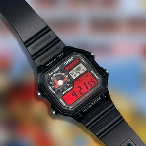 May include: Black Casio digital watch with a rectangular face and a black band. The watch face displays the time in red digital numbers, along with other functions. The watch has the text "CASIO", "WORLD TIME", and "ILLUMINATOR".