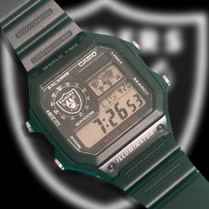Könnte beinhalten: Eine schwarze digitale Casio-Armbanduhr mit einem Raiders-Logo auf dem runden Zifferblatt, einer Weltkarte und den Aufschriften World Time, 5 Alarms, WR100M und 10 Year Battery. Die Uhr hat ein schwarzes Kunstharzarmband und ein Display.