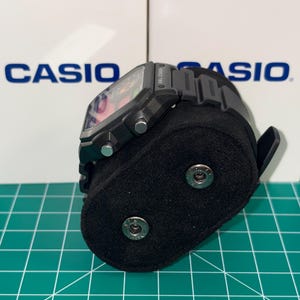 Puede incluir: Un reloj digital negro con una esfera cuadrada y una correa negra. El reloj se exhibe sobre un soporte de terciopelo negro. El nombre de la marca "CASIO" es visible en el fondo.