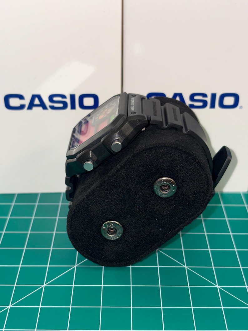 Puede incluir: Un reloj digital negro con una esfera cuadrada y una correa negra. El reloj se exhibe sobre un soporte de terciopelo negro. El nombre de la marca "CASIO" es visible en el fondo.