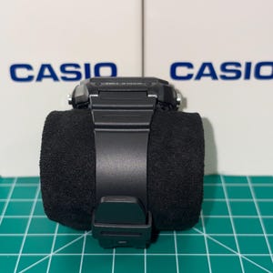 Peut inclure: Une montre num&eacute;rique Casio noire avec un bracelet noir est pr&eacute;sent&eacute;e sur un coussin cylindrique noir. La montre a un cadran carr&eacute; et une fermeture &agrave; boucle. Le mot "CASIO" est visible en arri&egrave;re-plan.